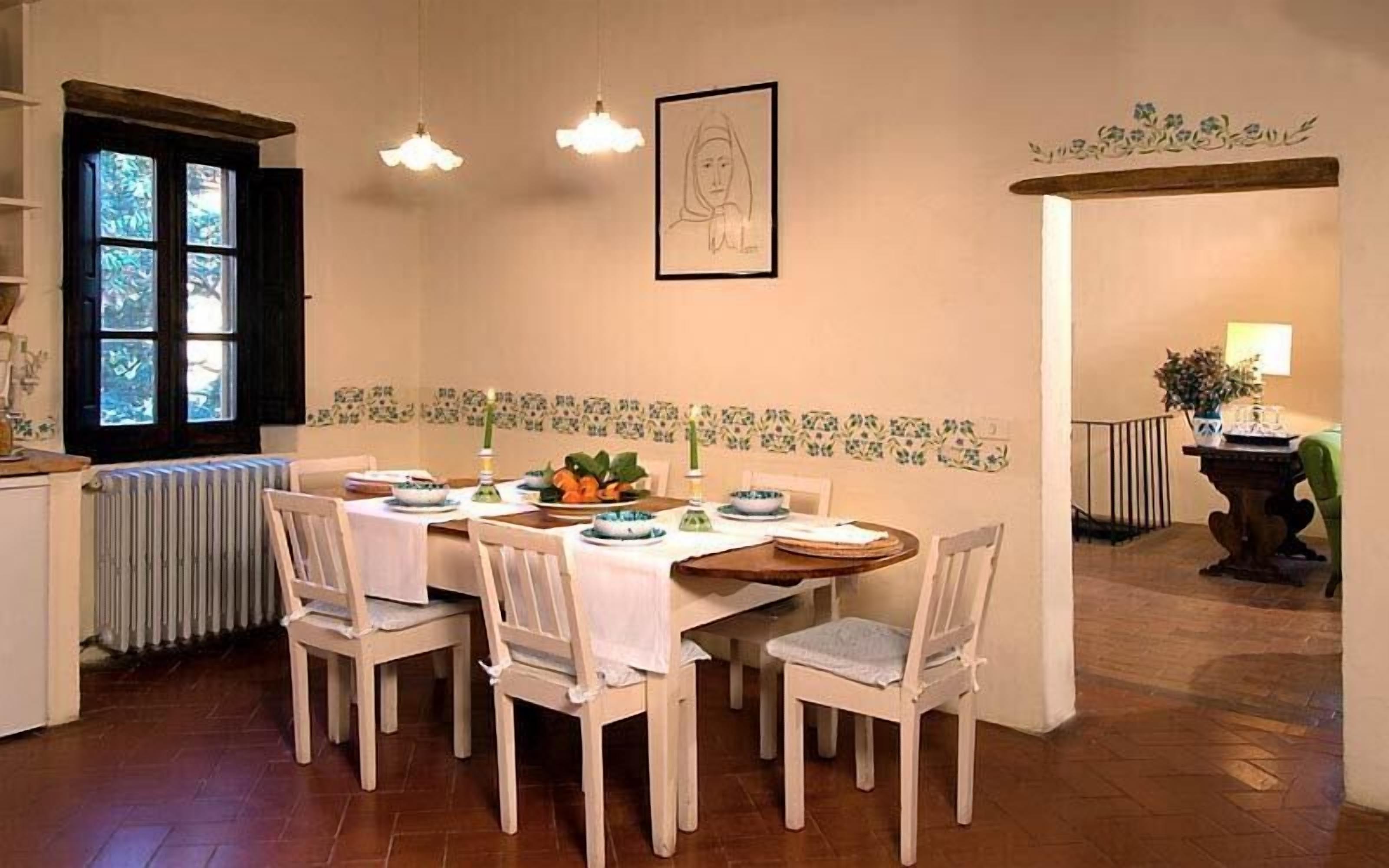 Ein Essbereich mit einem Tisch, gedeckt für sechs Personen, umgeben von weißen Stühlen. Links befindet sich ein Heizkörper, ein Fenster mit Jalousien und ein gerahmtes Bild an der Wand.