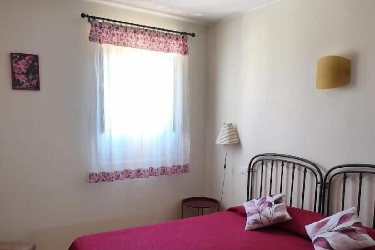 A bedroom with a bed, a window with a floral curtain, a bedside lamp, and a small stool.