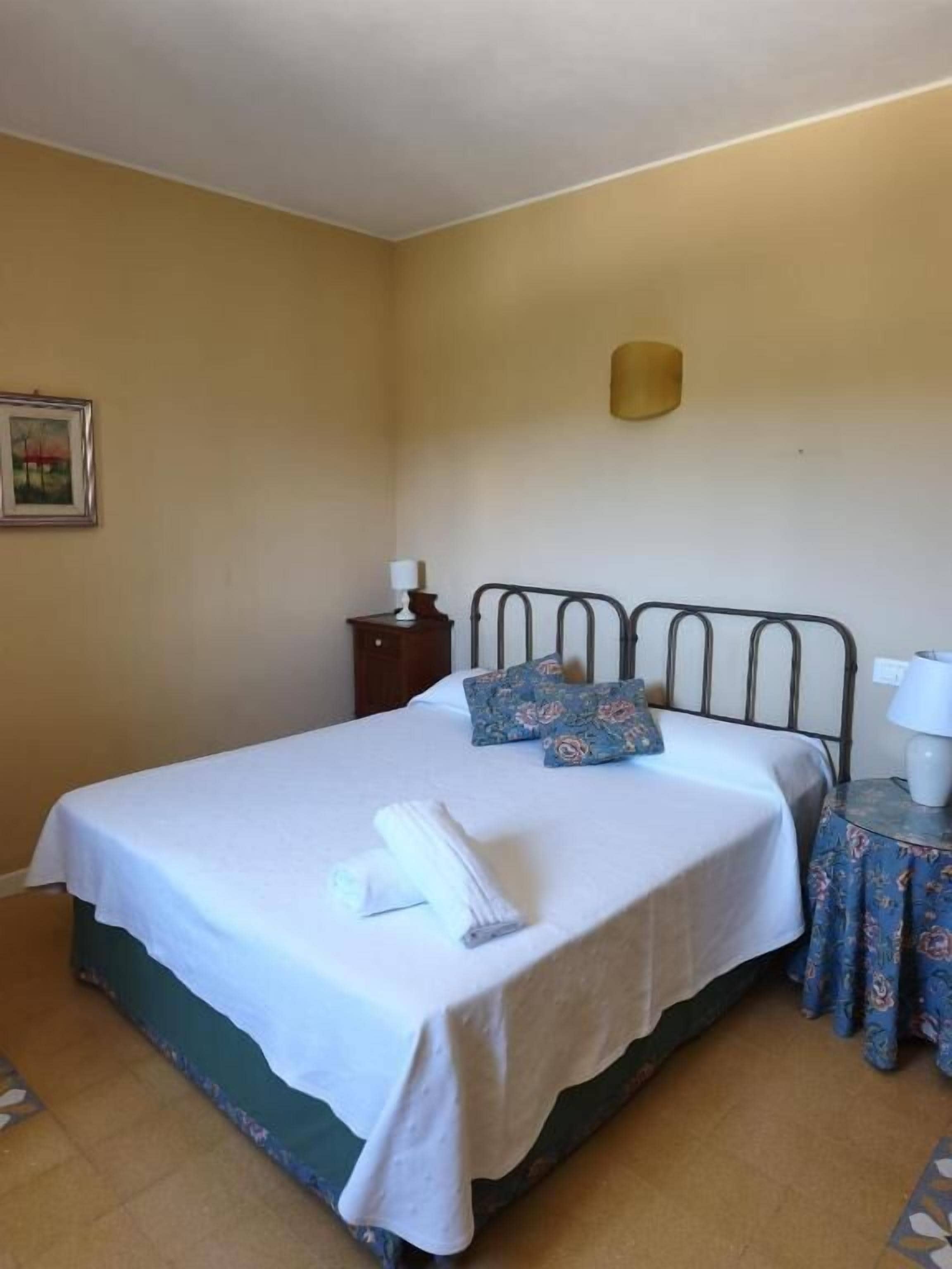 Una cama individual con una colcha blanca, dos almohadas, una mesita de noche con una lámpara y una pequeña mesa con mantel floreado.