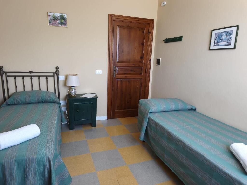 Una habitación con dos camas individuales, una puerta de madera, una mesita de noche pequeña con lámpara y suelo a cuadros.