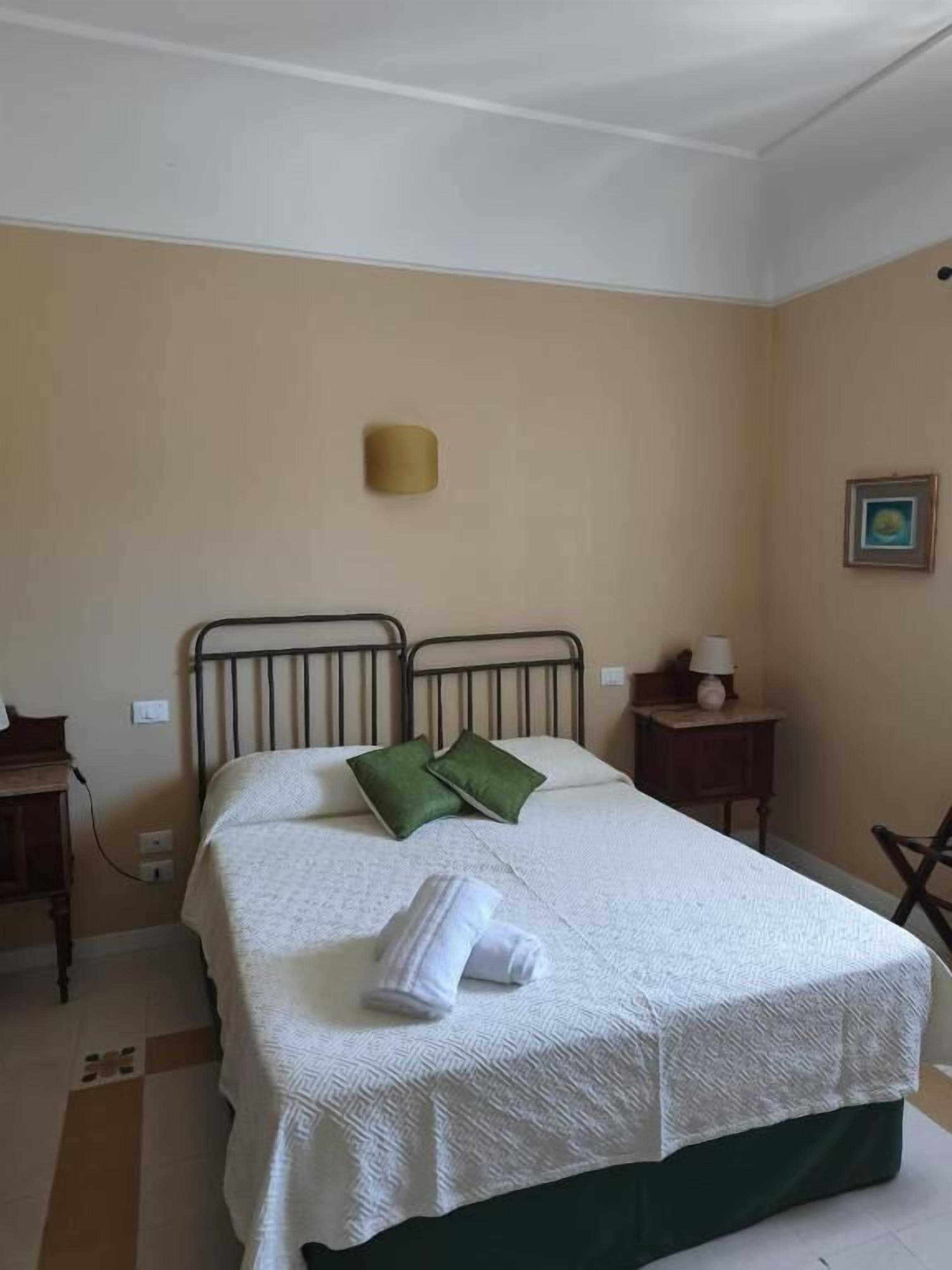 A single bed with a white bedspread and two green pillows, a bedside table with a lamp, and a wall-mounted light fixture.