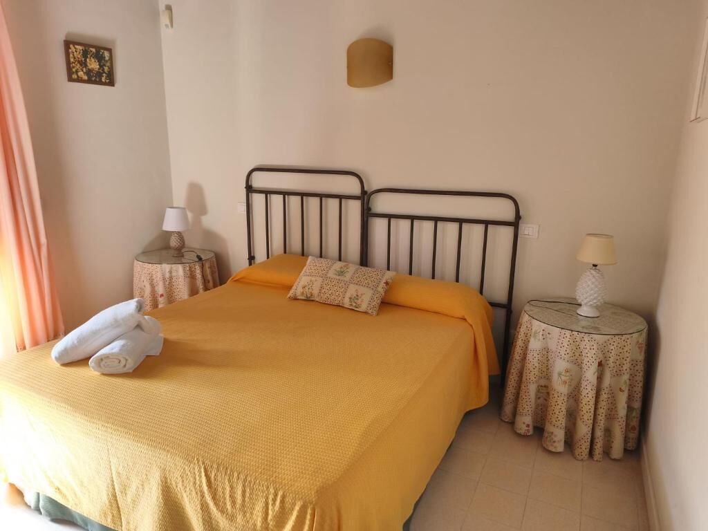 Una cama con una colcha amarilla, dos almohadas y una toalla enrollada al lado. Hay dos mesitas de noche con lámparas y una luz en la pared.