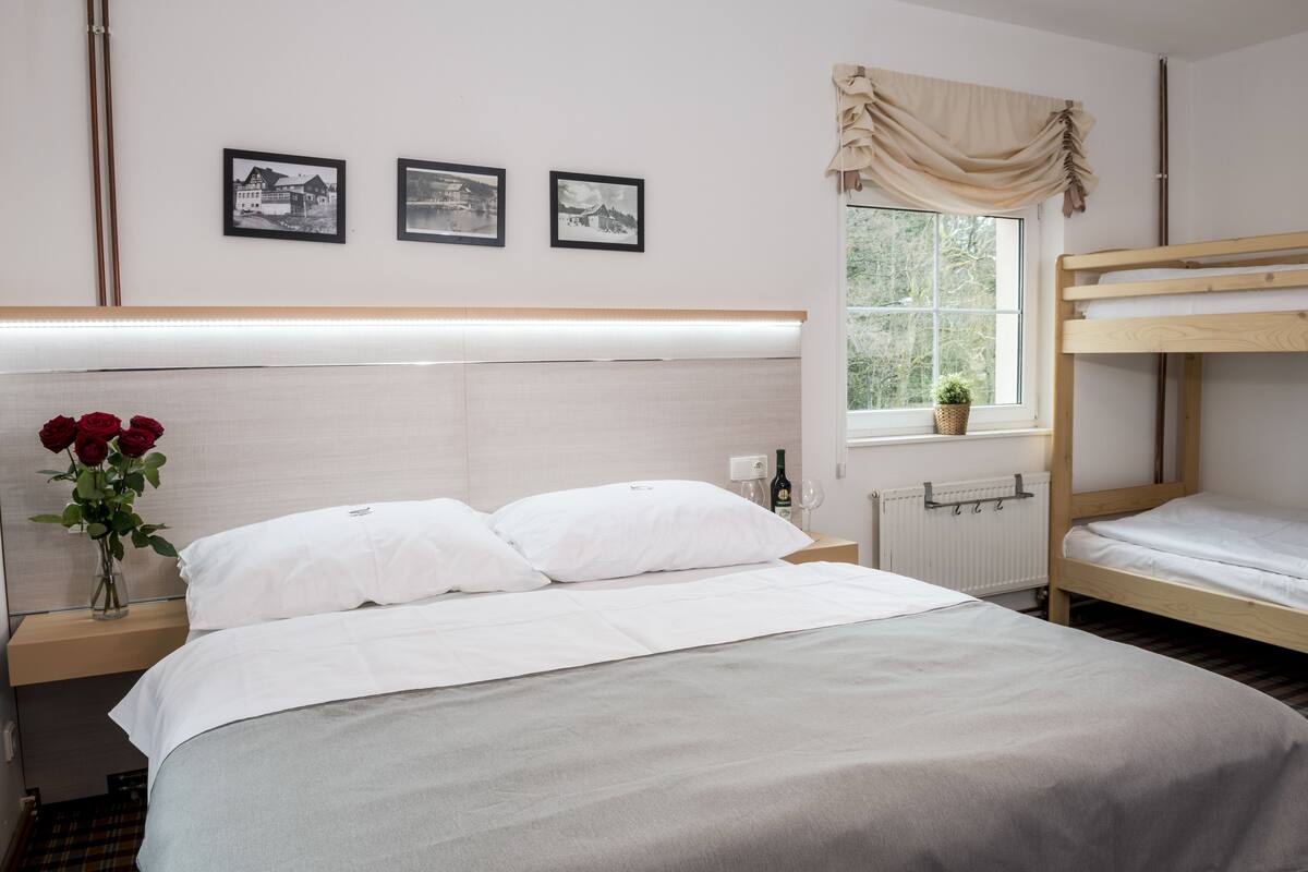 A hotel room with a large bed, a bedside table with a vase of red roses, a window with a view of greenery, and three black-and-white photographs on the wall.