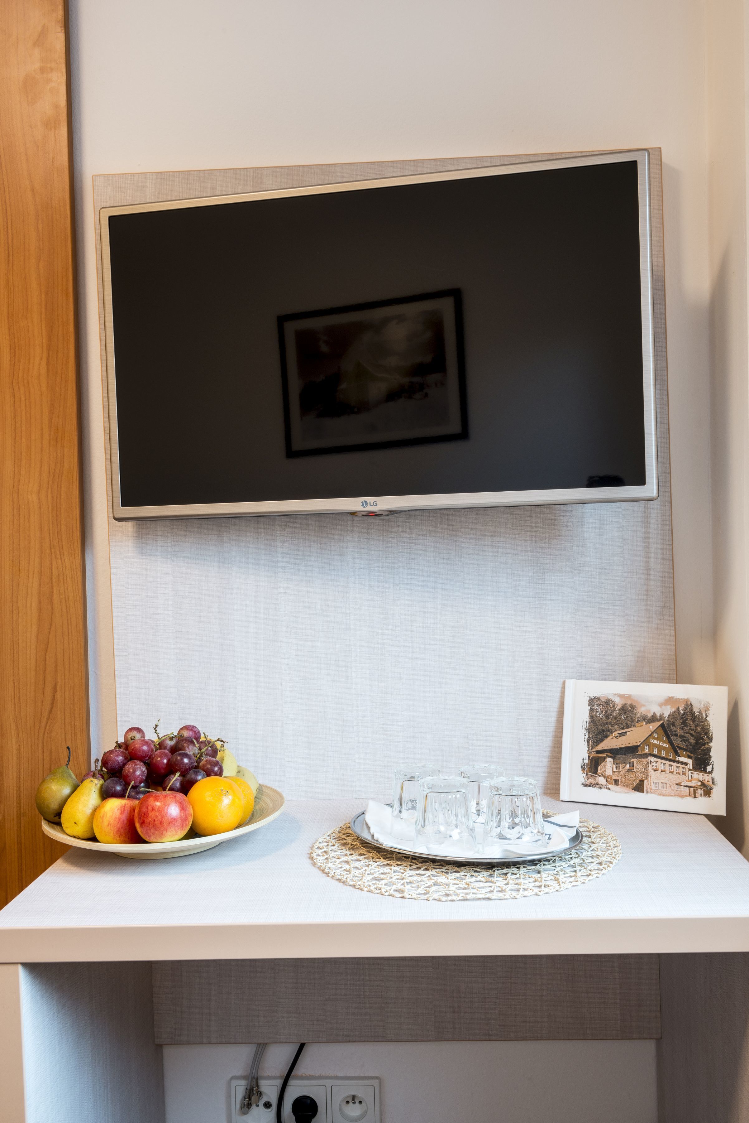 A flat-screen TV mounted on a wall, a fruit bowl with grapes and apples, a tray with glasses, and a framed picture.