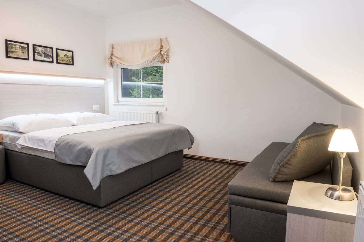 A bedroom with a bed, a nightstand, a lamp, a window with curtains, and a patterned floor.
