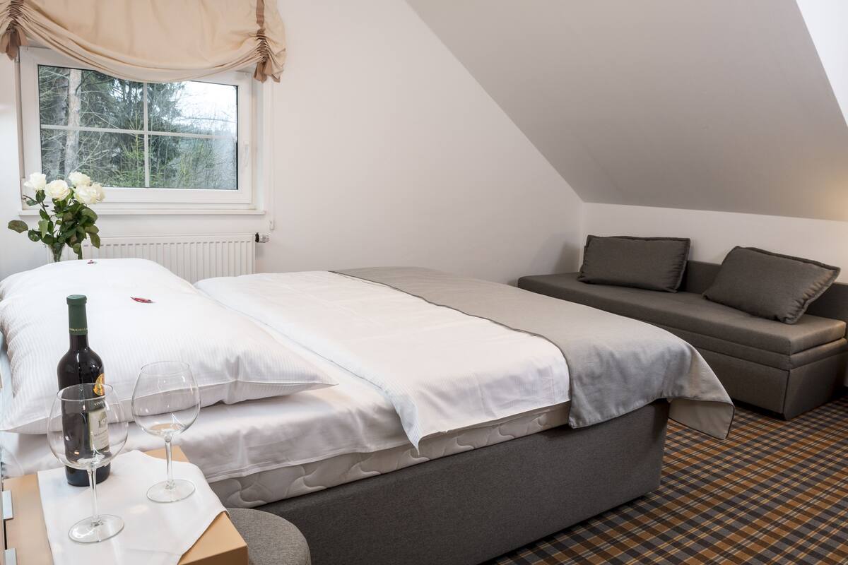 A bedroom with a bed, a sofa, a window with curtains, a radiator, and a bottle of wine on a table.