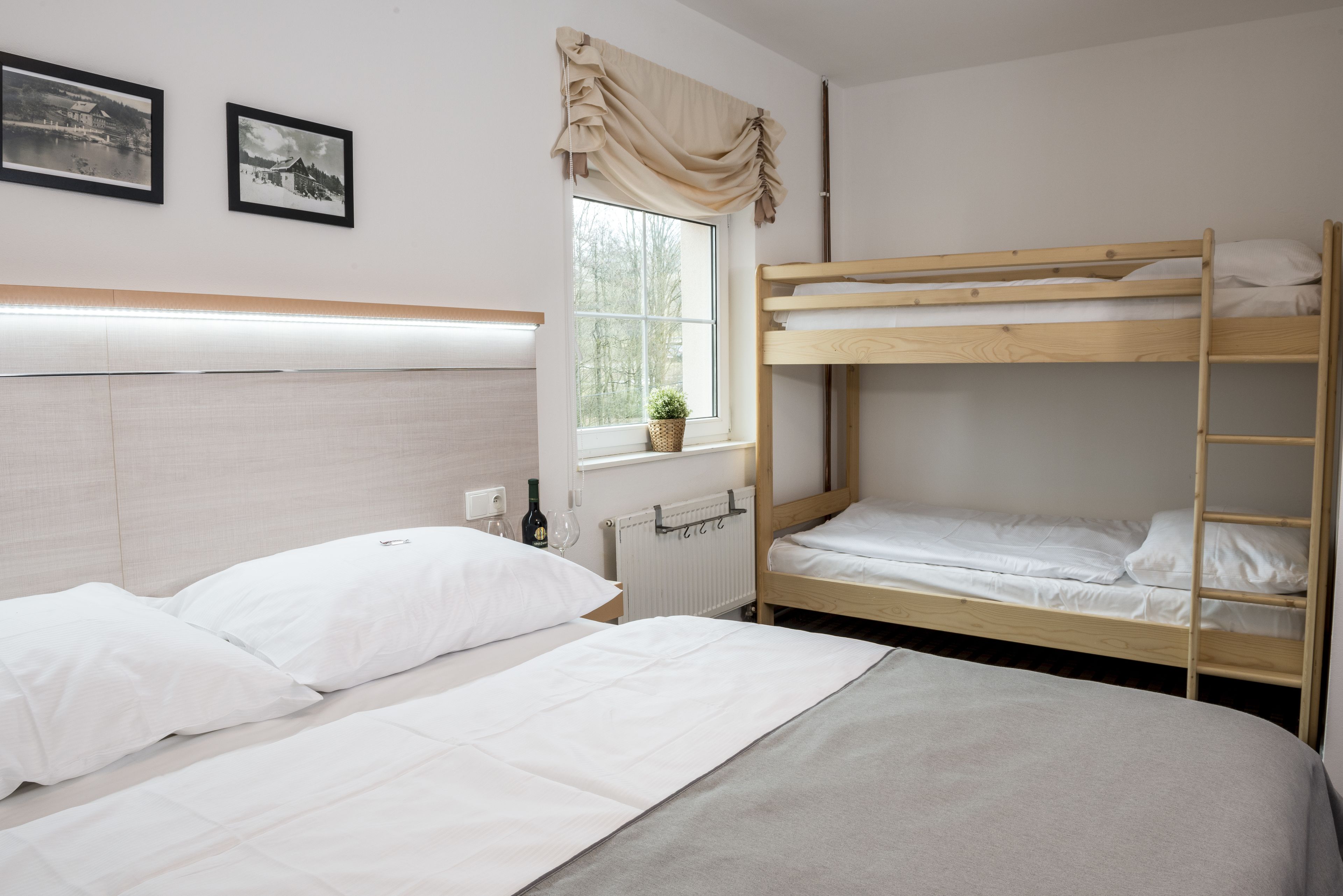 A room with a bunk bed, a single bed, a window with curtains, a radiator, and two framed pictures on the wall.