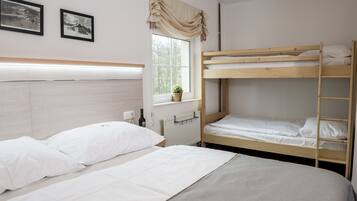 Una habitación con una cama litera, una cama individual, una ventana con cortinas, un radiador y dos cuadros enmarcados en la pared.