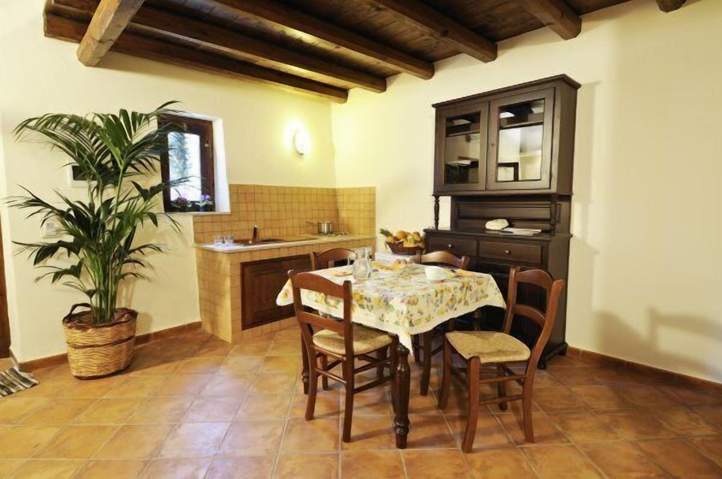 A dining area with a table set for four, surrounded by wooden chairs. A potted plant is on the left, and a cabinet is against the wall.