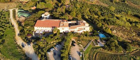 An aerial view of a large estate with multiple buildings, including a main house with a tiled roof, surrounded by lush greenery and vineyards.