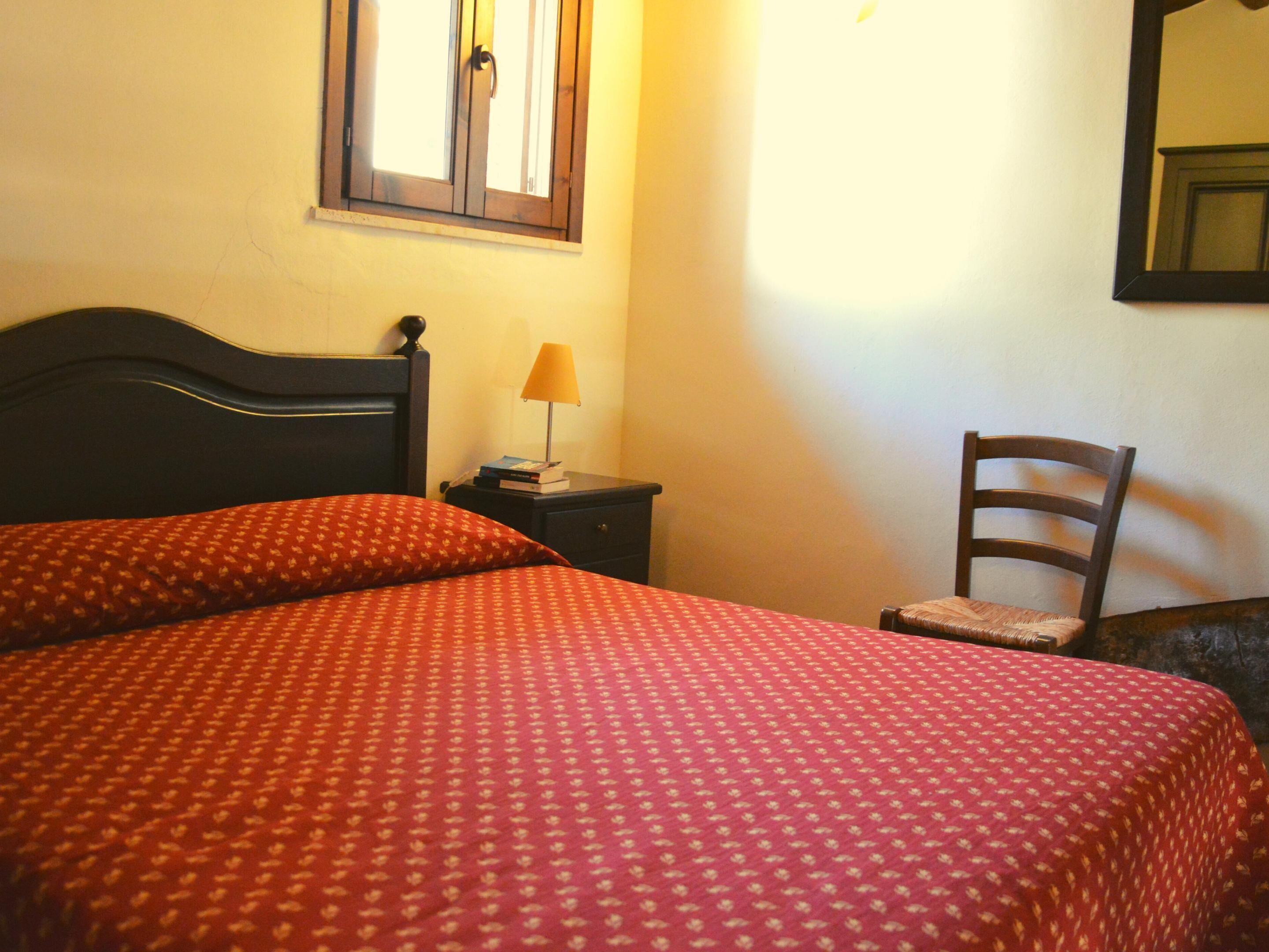 Cama bem arrumada com colcha vermelha estampada, mesa de cabeceira com abajur e uma cadeira em quarto com janela e espelho na parede.