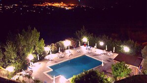 Uma piscina ao ar livre com espreguiçadeiras e guarda-sóis, rodeada por árvores e vista para um horizonte urbano à noite.