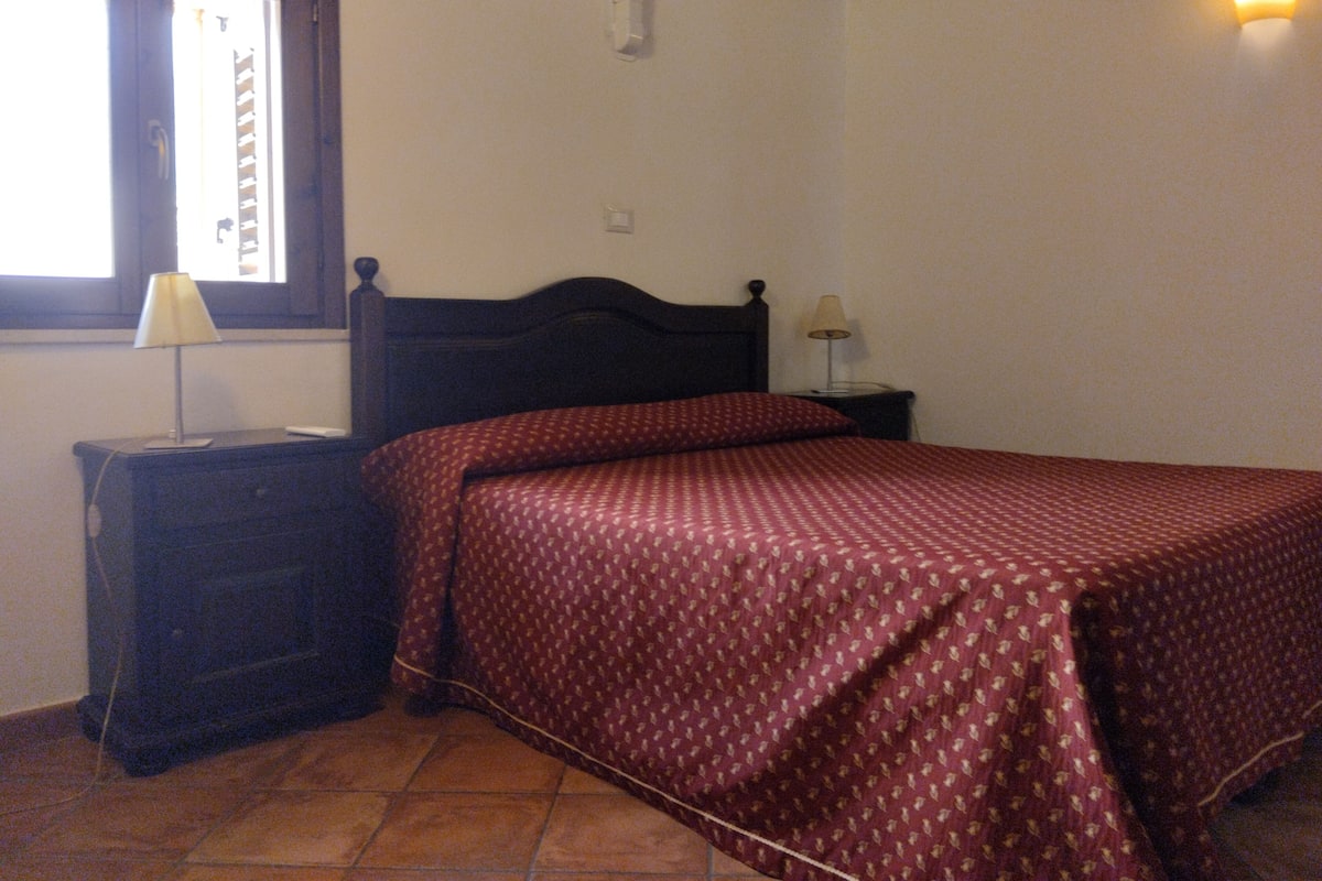 Ein Schlafzimmer mit einem Bett, einem Nachttisch, einer Lampe, einem Fenster und einer Wandleuchte.