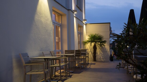 An outdoor seating area with tables and chairs, a palm tree, and a building with large windows and a sign that reads "HOTEL". - Goethe Conference Hotel by Trip Inn (Frankfurt)