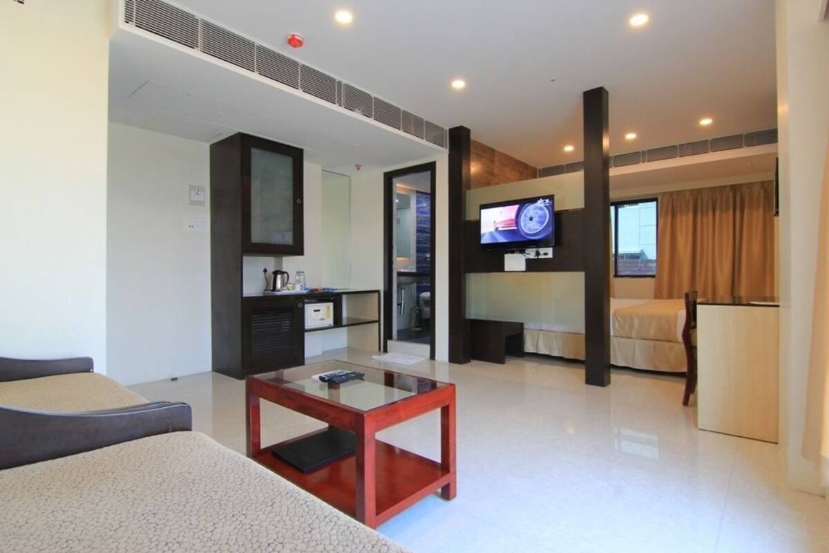 A modern hotel room with a glass coffee table, a flat-screen TV, a bed, and a kitchenette.
