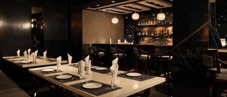 Un restaurante con largas mesas rectangulares dispuestas para comer, una zona de bar con estantes llenos de botellas y un diseño moderno de techo.