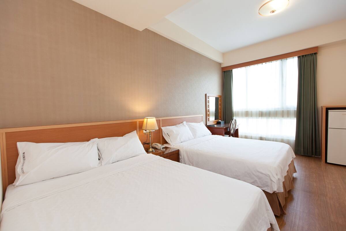 Habitación de hotel con dos camas, una mesita de noche con lámpara, un escritorio y una ventana con cortinas.