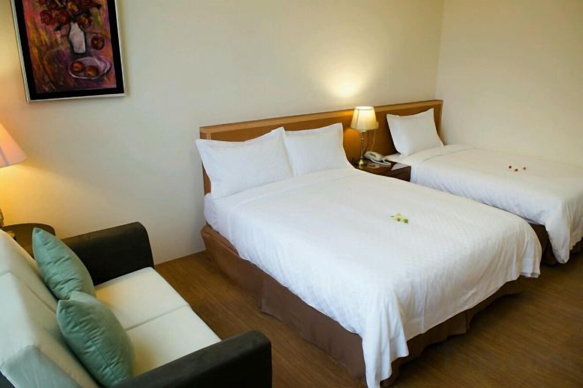 A hotel room with two single beds, a sofa, and a nightstand with a lamp.