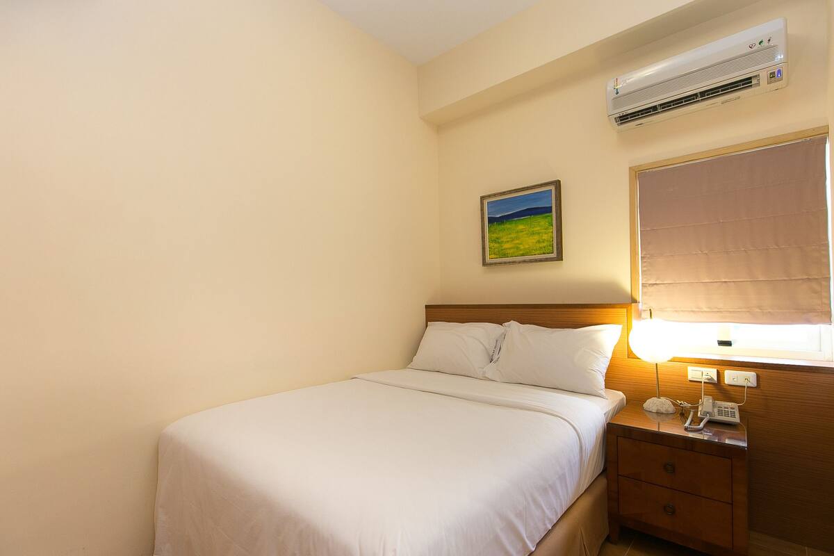 Una habitación de hotel con una cama, una mesita de noche con una lámpara, aire acondicionado, una ventana con persiana y un cuadro en la pared.