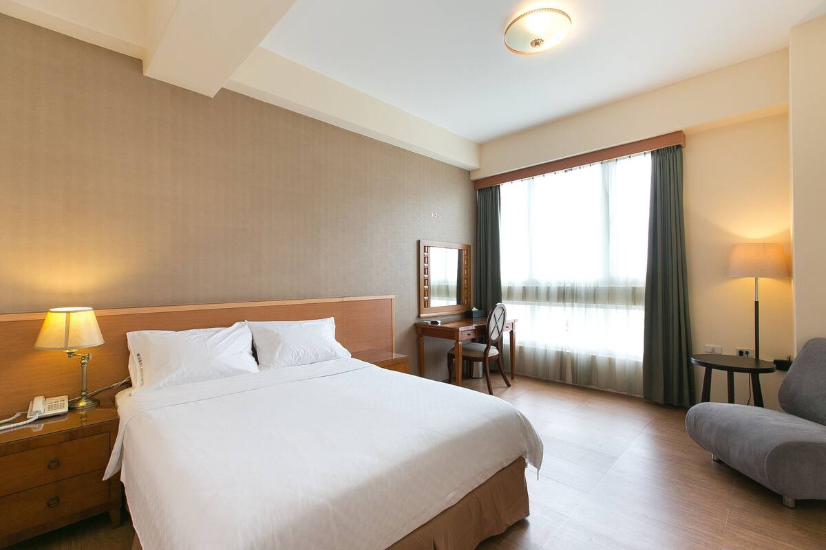 Una habitación de hotel con una cama grande, un escritorio, una silla, una lámpara y una ventana con cortinas.