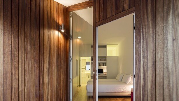 A room with wooden panel walls and a sliding door leading to a bedroom with a bed and a desk.