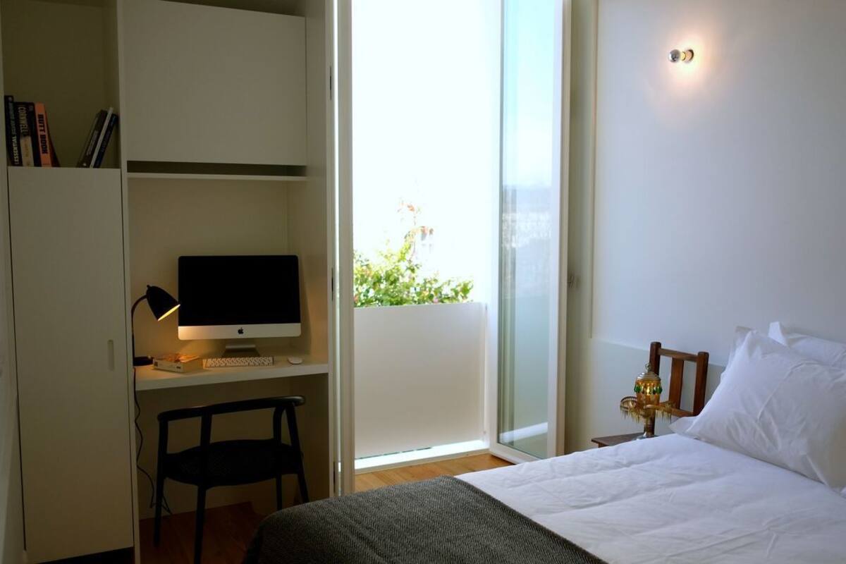A bedroom with a bed, a desk with a computer, a chair, and a window with a view of greenery.