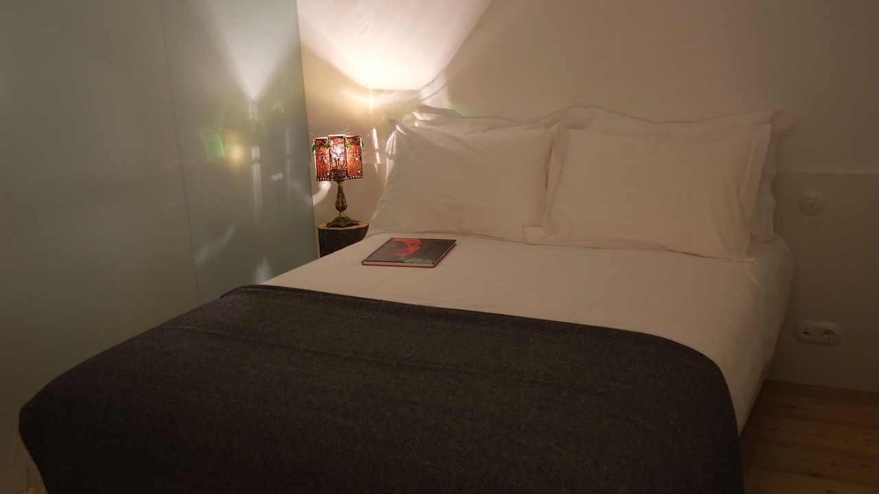 A neatly made bed with white linens and a dark throw blanket, a bedside lamp with a patterned shade, and a book on the bed.