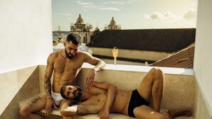 Two men on a rooftop terrace with a cityscape view, one sitting and the other standing.