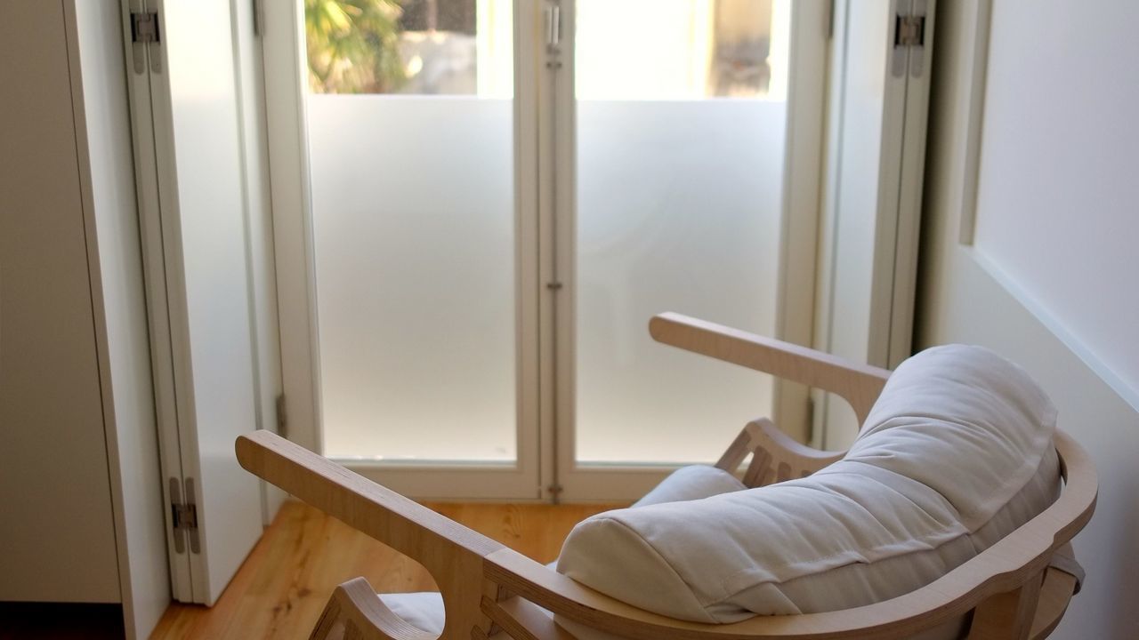 Una cadira bressol de fusta amb coixí blanc a una habitació amb una finestra gran i una porta.