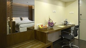 Une chambre d’hôtel avec un bureau en bois, une chaise, un lit avec une tête de lit, un miroir et un petit vase avec des fleurs.