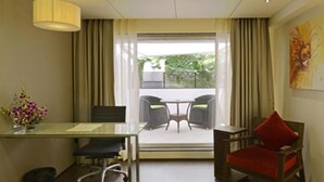 Une chambre d’hôtel avec un bureau, deux chaises, dont une rouge, une petite table, une lampe, un tableau et une vue sur la verdure à l’extérieur.