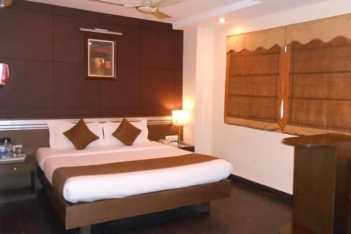 A hotel room with a large bed, a desk, a chair, a window with curtains, a framed picture on the wall, and a ceiling fan.