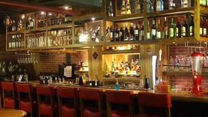 A bar area with multiple shelves of bottles, a counter with a cash register, and red chairs. - Hotel O2 VIP (Barasat)