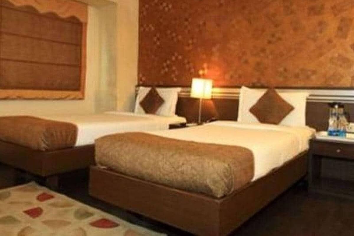 A hotel room with two beds, a wooden headboard, a nightstand with a lamp, and a patterned rug.