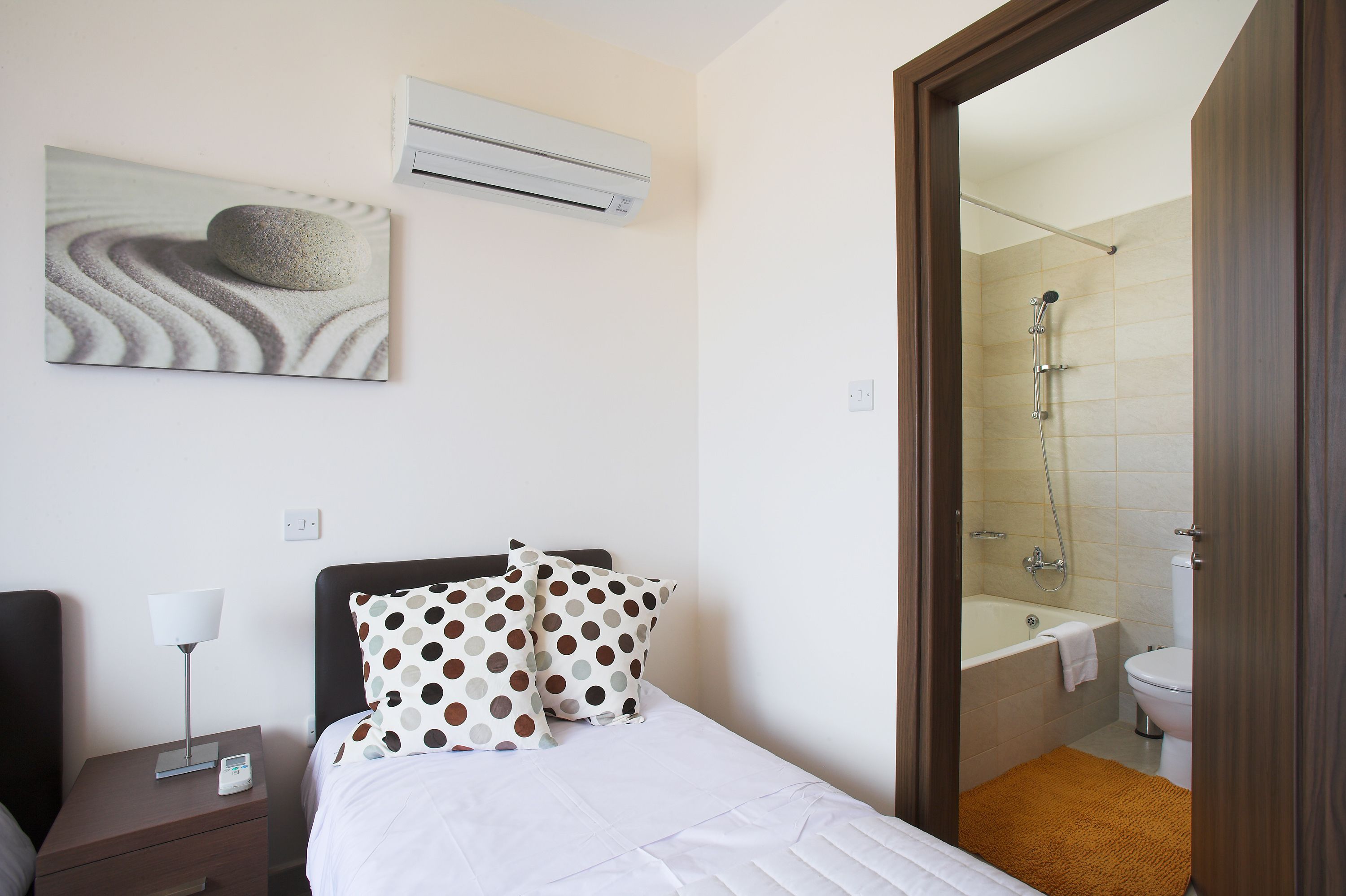 A hotel room with a bed, a nightstand, a wall-mounted air conditioner, and a bathroom visible through an open door.