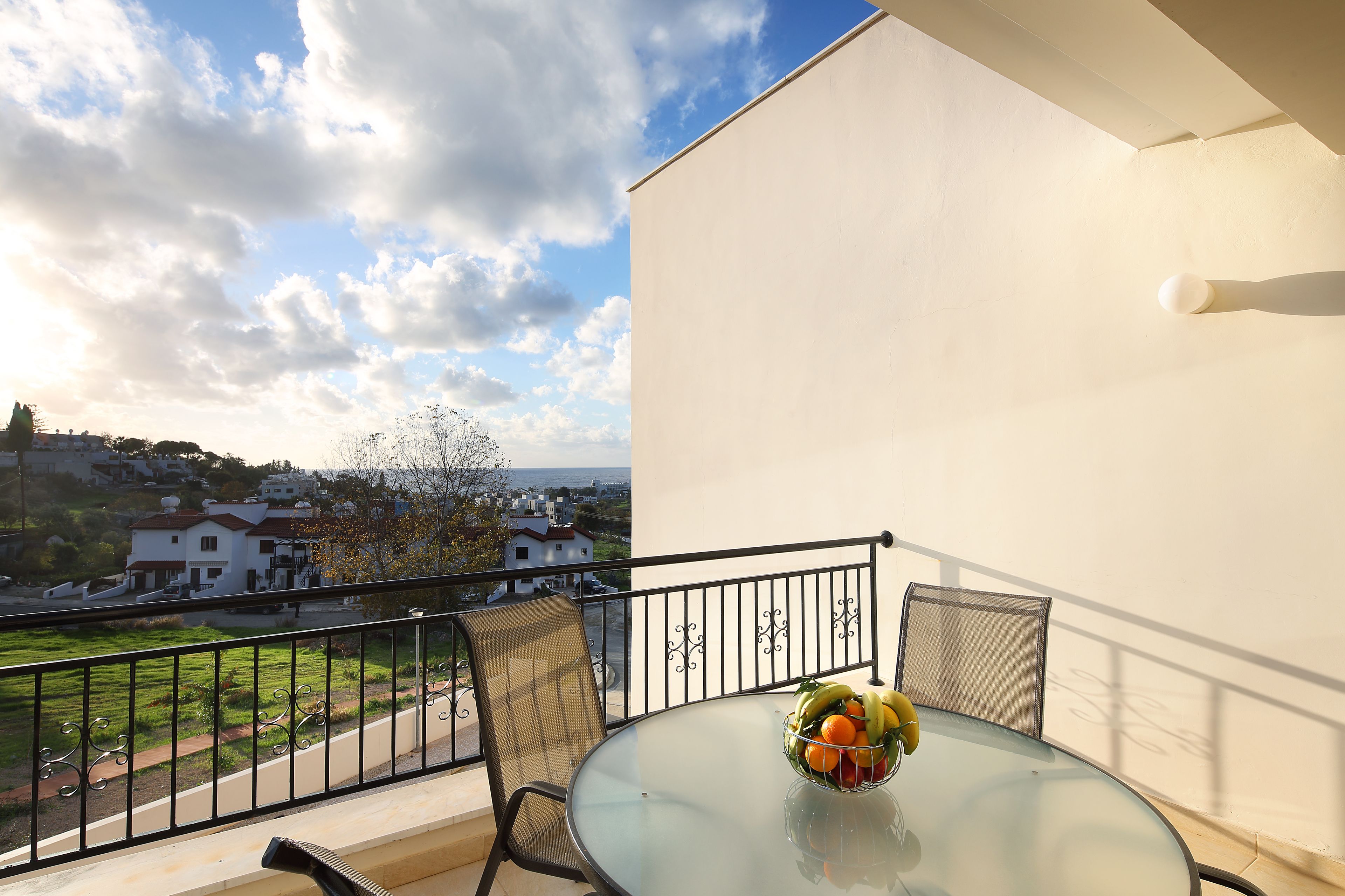 A balcony with a glass table and a bowl of fruit, overlooking a residential area with houses and greenery.
