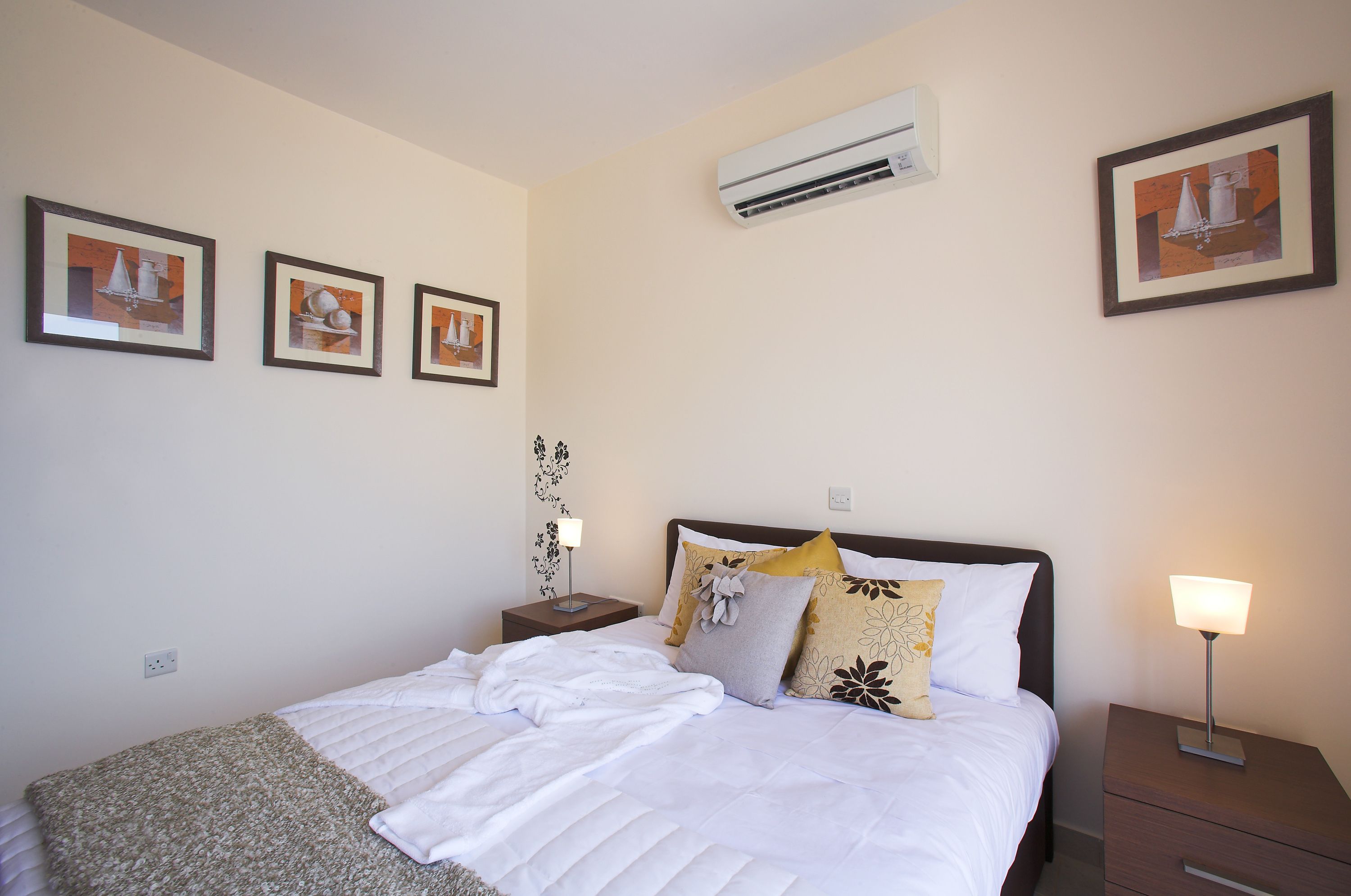 A neatly made bed with white linens, a bedside table with a lamp, and three framed pictures on the wall.