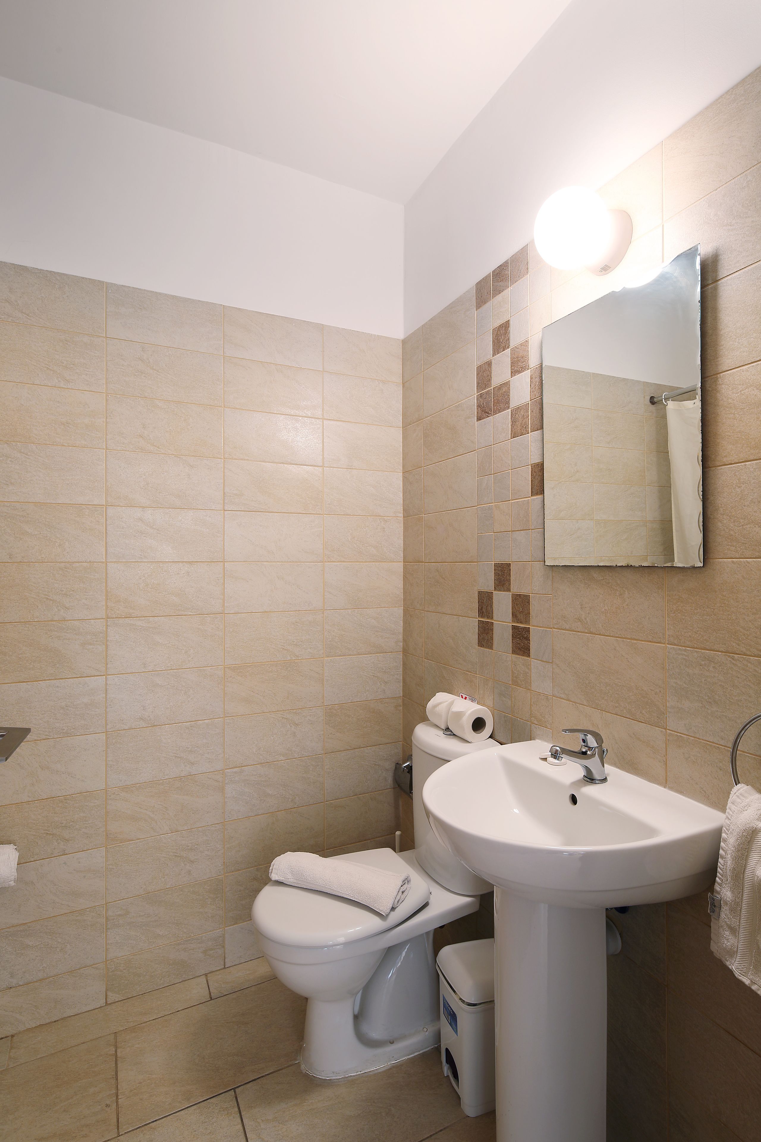 A bathroom with beige tiles, a white sink, a toilet, and a mirror with a light.