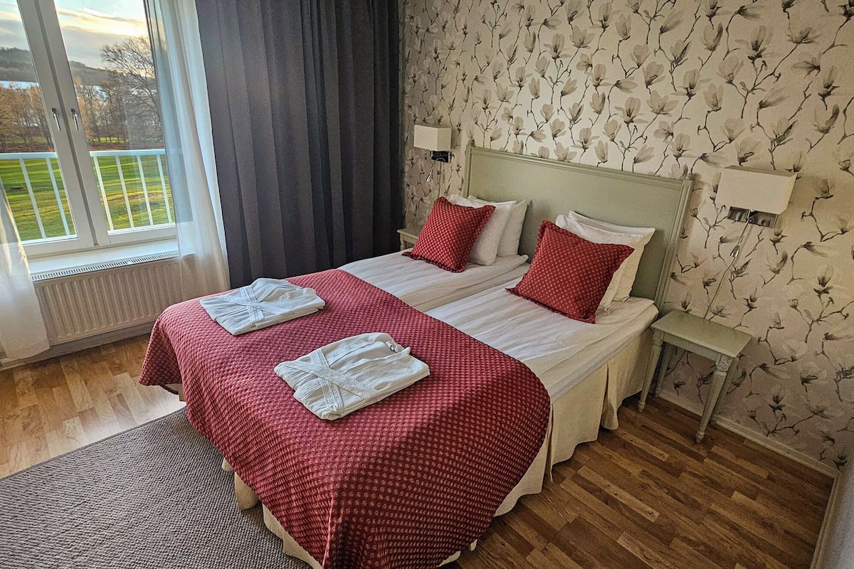 Cama doble con ropa de cama roja y blanca, una mesita de noche, una lámpara y vistas a un paisaje verde a través de una ventana.