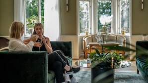 Dos personas sentadas en sofás verdes en una habitación con ventanas amplias, una mesa de centro redonda de vidrio y una mesita auxiliar de madera con una lámpara.