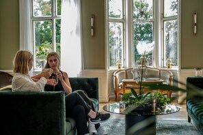 Dos personas sentadas en sofás verdes en una habitación con ventanas amplias, una mesa de centro redonda de vidrio y una mesita auxiliar de madera con una lámpara.