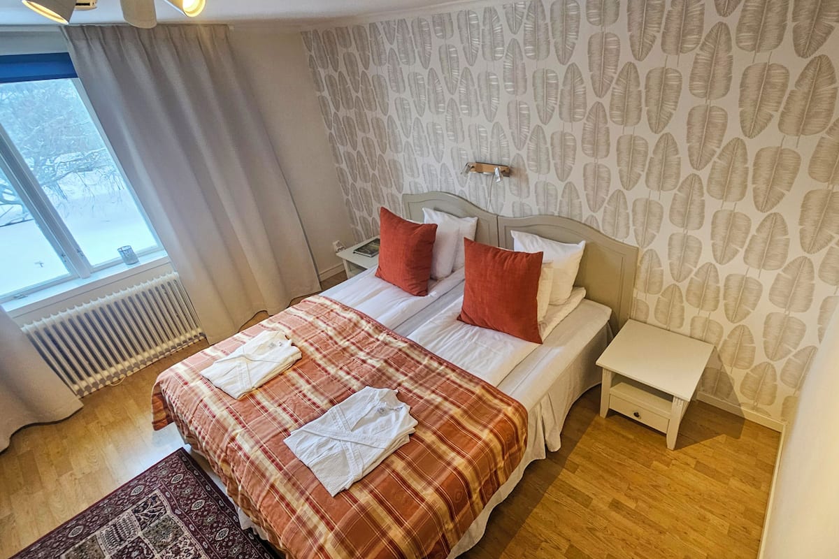 Habitación de hotel con cama, dos mesitas de noche, un radiador, una ventana con cortinas y una pared con diseño.