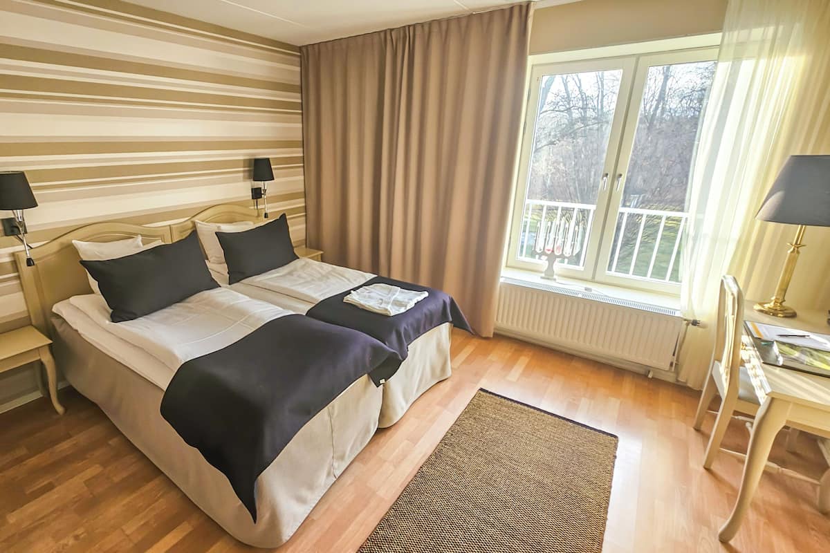 Cama doble con cabecera, mesitas de noche, una ventana con cortinas y vistas a árboles.
