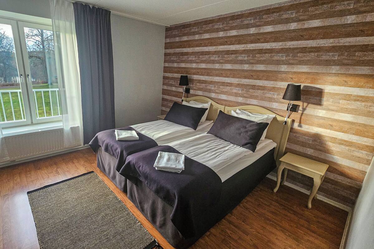 Un dormitorio con dos camas, una ventana con cortinas, piso de madera, una alfombra y un panel decorativo de madera en la pared.