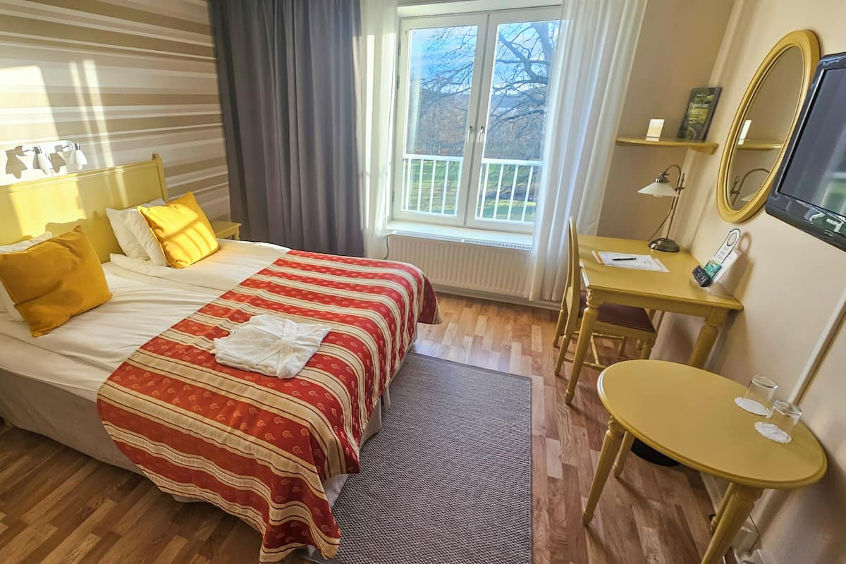 Habitación de hotel con cama, mesita de noche, silla, escritorio, espejo y ventana con vistas.