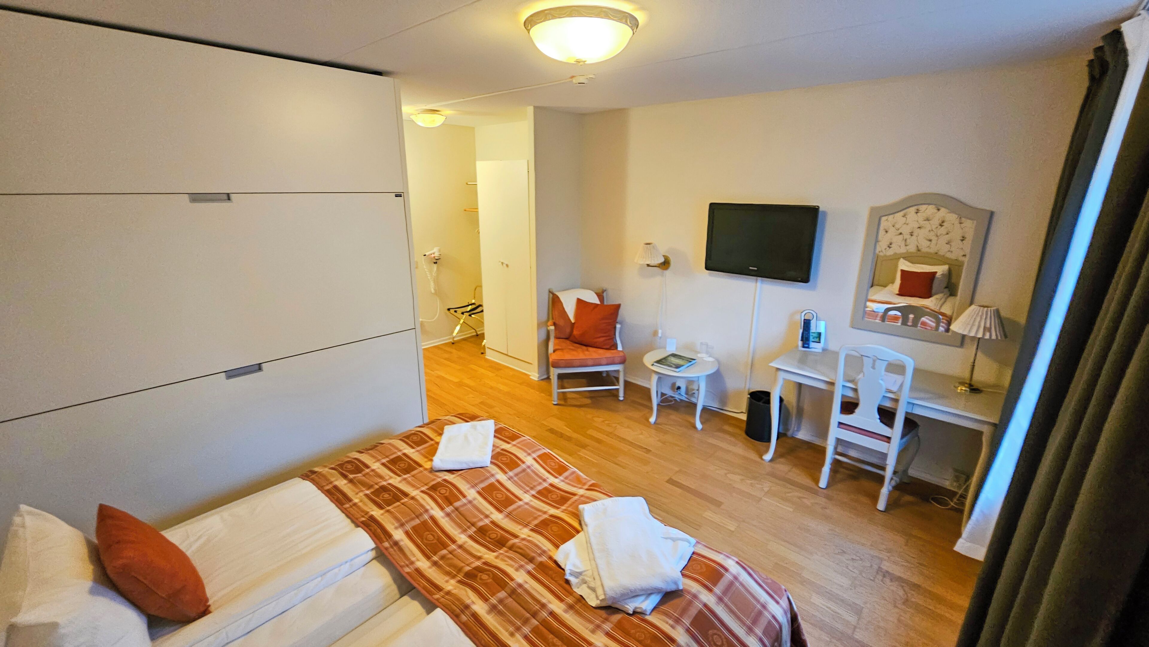 Quarto de hotel com cama, escrivaninha, poltrona, televisão e guarda-roupa.