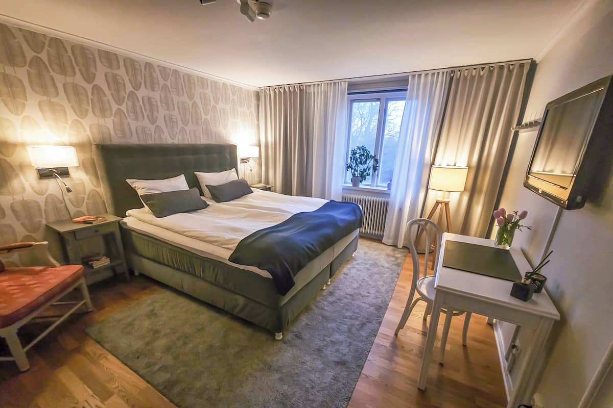 Habitación de hotel con cama, escritorio, silla, ventana con cortinas y una planta.