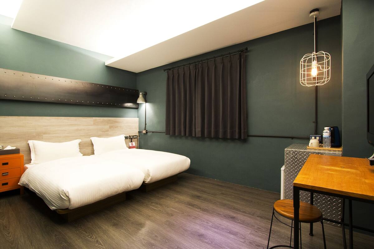 Ein modernes Hotelzimmer mit einem Holz-Kopfteil, einem Bett mit weißen Leinen, einem Schreibtisch mit Stuhl, einer Pendelleuchte und einem Fenster mit Vorhängen.
