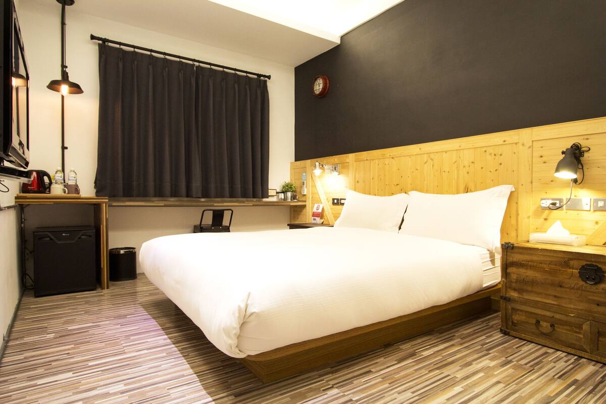 Ein Hotelzimmer mit einem großen Bett, einem Holz-Kopfteil, einem kleinen Schreibtisch mit Lampe, einem Fenster mit Vorhängen und einer Wanduhr.