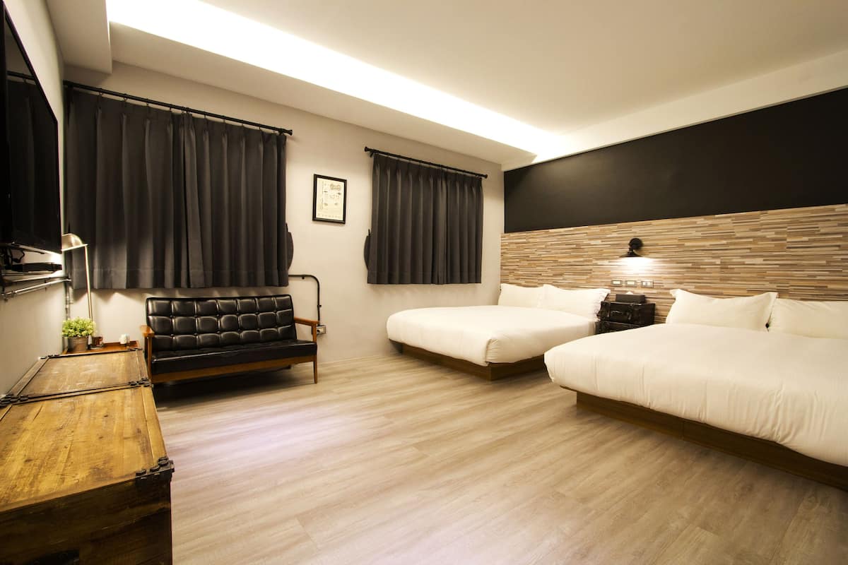 Ein modernes Hotelzimmer mit Holzboden, einer Couch, zwei Einzelbetten, einem Holzkopfende und einem kleinen Tisch mit einer Lampe.
